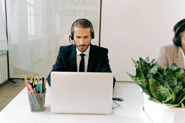 Focused customer support agent using laptop and headset in a modern office setting with greenery. Comparing per-agent and usage-based support pricing models