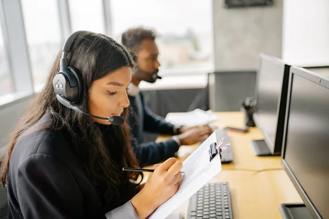 Two call center agents wearing headsets working at desks with computers in a modern office. Measuring whether quality reviews improve customer outcomes